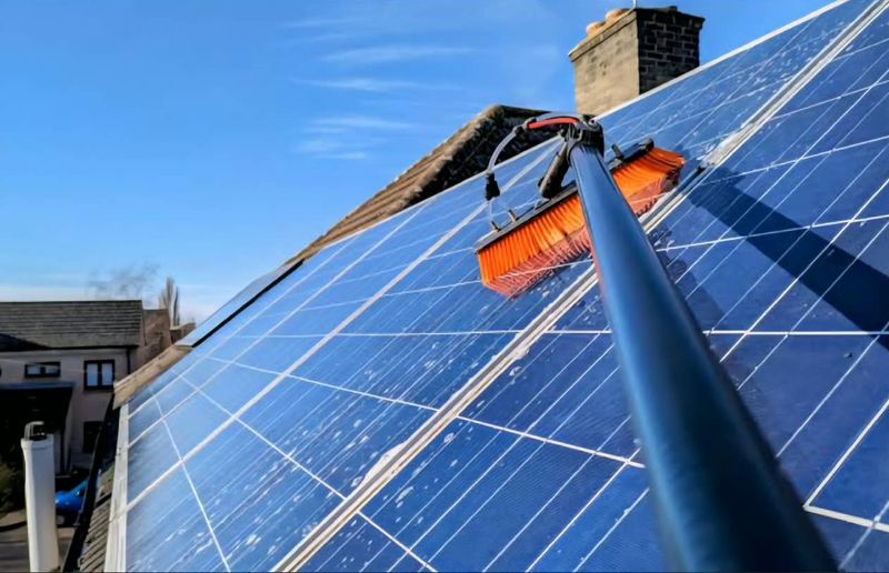 Close-up of an orange soft-bristle brush on a water-fed pole cleaning a pitched-roof solar array under a blue sky